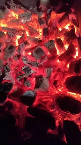 A close-up shot focuses on glowing red embers and charcoal inside a barbecue grill. The camera slowly pans downwards, revealing the metal structure of the grill and a grassy area below. There are no people or animals visible. The lighting suggests it is dusk or nighttime, with the embers providing the primary light source.