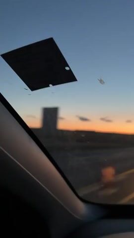 A first-person view from inside a vehicle shows a highway at dusk. The sky is a deep blue with streaks of orange and pink near the horizon. The road ahead is visible, with yellow lane dividers and illuminated construction cones. There are a few scattered clouds in the sky. On the left side of the windshield, a black square object is attached, possibly a toll transponder or registration sticker. There are a few smudges or streaks on the windshield. The vehicle appears to be in motion, indicated by the blur of the scenery and the perspective of the road. The overall environment is open and appears to be a rural or semi-rural highway.