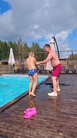 A boy in blue patterned swim trunks, wearing goggles, stands on the edge of a wooden deck next to a swimming pool. A man in red shorts and white shoes stands behind him, with his hand on the boy's back. The boy jumps into the pool, creating a splash. Another person is visible in the pool. Pink flip-flops are on the wooden deck. The background shows trees, a fence, lounge chairs, and umbrellas.