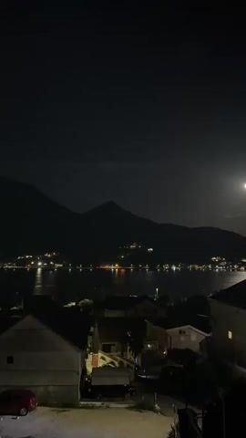 The video shows a nighttime view overlooking a body of water and a hillside town illuminated by artificial lights. A full moon is visible in the upper right corner of the frame. In the foreground, there are houses with lights on, and a red car is parked on the left. The water reflects the lights from the town, creating a shimmering effect. The overall scene depicts a peaceful, nocturnal landscape.