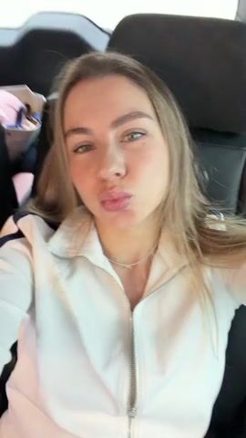 A young woman with blonde hair and blue eyes makes kissing faces at the camera. She is sitting in the passenger seat of a car, wearing a white jacket and a necklace. The car appears to be in motion, with a blurred green landscape visible through the window. The woman winks at the camera in the final frame.
