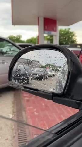 The video opens with a close-up of a car's side mirror, its surface covered in raindrops. Reflected in the mirror is a scene at a gas station, with several cars lined up. The overall mood is somewhat dreary due to the rain.

As the video progresses, the camera pans slightly, revealing more of the gas station environment. We see gas pumps, one of which has the number "5" prominently displayed on its red and white structure. There are also other cars waiting or being serviced. A person in a bright orange vest, likely a gas station attendant, is visible near a pump. The floor of the gas station is paved with red bricks. The sky appears overcast, suggesting a cloudy or rainy day.

The scene is set in Ladozhskaya, Russia, as indicated by the context. The visible text on the gas pump includes the number "5" and what appear to be prices or fuel types like "95" and "92". The activity taking place is a common one at a gas station: vehicles refueling. The presence of raindrops on the mirror and the overcast sky indicate that it is likely daytime and raining. The overall impression is a mundane, everyday scene at a gas station under inclement weather.