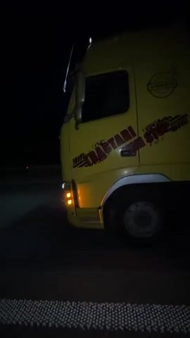 A yellow truck is parked at night on the side of a road, next to a metal barrier. The truck has "VOLVO" and "TRAATARI MA STOP" written on its side. Another red truck is visible in the distance, facing the yellow truck. The yellow truck's side lights are on, illuminating the ground around it. The video pans from the front of the yellow truck to its rear, then back to the front, revealing the red truck in the distance. The metal barrier extends along the side of the road, separating the trucks from the other side of the road. The red truck's headlights are on, casting a bright light on the road ahead. The scene is dark, with only the truck lights and some streetlights providing illumination.