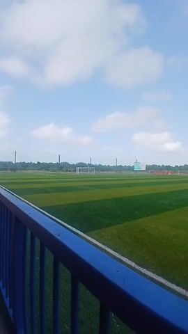 Here's a concise description of the scene:

The video shows a view from behind a blue railing of a soccer field. The field has green and yellow artificial turf. A white line marks the edge of the field closest to the camera. In the distance, people in red jerseys are visible on the field. A soccer ball sits near the railing. The sky is blue with white clouds. Beyond the field, there are trees and possibly buildings.