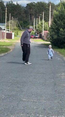 A short outdoor video shows a baby walking towards an adult male on an asphalt road. The baby is wearing a gray beanie, gray overalls, and black and white shoes. The baby appears to be around one year old. The man is wearing a gray sweatshirt, black and white patterned pants, white sneakers, and a gray baseball cap. He is standing still, facing the baby with his arms outstretched. The road is surrounded by houses, trees, and telephone poles. The sky is partly cloudy. The video is taken during the day. The camera angle is straight on, showing both the baby and the man in full view.