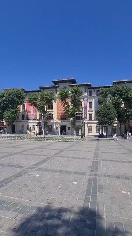 A wide shot shows a large, multi-story building with banners hanging on its facade, set against a bright blue sky. The building is constructed of light-colored stone and features multiple windows and balconies. In front of the building is a large, paved plaza with a checkered pattern of stones. Several trees with green foliage are planted in front of the building, partially obscuring it. The plaza appears to be mostly empty, with a few people visible in the distance, walking near the building. The lighting suggests it is a sunny day.