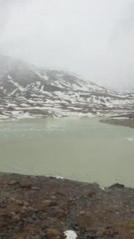 The video depicts a view from inside a train, looking out the window. Raindrops are visible on the glass. Outside, there is a lake with a milky green color, likely due to glacial meltwater. In the background, a mountain range is partially covered in snow and obscured by dense fog or clouds. The foreground shows a grassy, rocky slope leading down to the lake. The overall atmosphere is cold and misty. There is some indiscernible indistinct chatter audible.