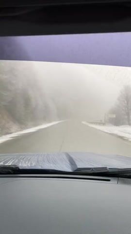 The video captures a first-person view from inside a vehicle driving along a paved road. The dashboard and windshield wipers are visible at the bottom of the frame. The road appears wet and slushy, with snow covering the shoulders and surrounding landscape.

Heavy fog or mist significantly reduces visibility, obscuring distant features of the mountainous terrain. On the left, a snow-covered rocky slope with sparse, frosted trees is visible. To the right, there's a snow-dusted roadside and a metal guardrail, with a faint, unreadable sign further in the distance. The vehicle is in motion, navigating through the challenging, low-visibility conditions typical of a cold, wintery environment. Audio of music is playing in the background. The location is Lago Escondido, Argentina.