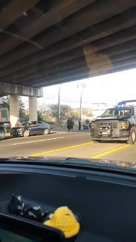 The video, filmed from the perspective of a car driver, depicts a traffic stop. Initially, a police truck, with blue and red lights activated, is seen. The camera pans right, revealing a line of police vehicles on the road. Several police officers, dressed in dark uniforms, are visible near the vehicles and crossing the road. In the background, there is a cityscape with buildings and a mountainous horizon. The sky is partly cloudy, and the sun casts shadows. The car's dashboard is visible at the bottom of the frame, featuring a yellow fuzzy object and a mounted tablet. The video captures the scene as the driver waits behind the police activity.
