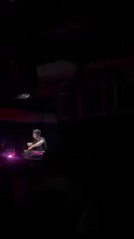The video shows a DJ performing in a dimly lit club. The DJ is standing behind a mixing console, manipulating controls with both hands. Several people are visible in the foreground, facing the DJ, presumably the audience. The club has a disco ball and large speakers above and behind the DJ. The lighting is predominantly purple.