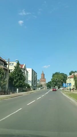 The video shows a view from a vehicle driving down a street in Gryfino, Poland. The scene is a sunny day with a clear blue sky and a few wispy clouds. The street is lined with buildings on both sides, with trees and greenery interspersed.

On the left side of the road, there are multi-story residential buildings. On the right, there are also buildings, and further down, a tall brick tower with a pointed roof is visible. This tower appears to be a significant landmark. Traffic lights are visible, indicating an intersection ahead. A few cars are on the road, including a red car in front of the camera's vehicle. Some pedestrians are also seen walking on the sidewalks.

The time of day appears to be daytime, likely late morning or afternoon, given the bright sunlight casting shadows. The weather is fair and pleasant. The overall atmosphere is that of a typical town street scene on a bright, clear day.

There is some text visible on road signs. On the right side of the road, there are blue road signs with white text and symbols, indicating directions and possibly speed limits or other traffic regulations. One sign shows a left-pointing arrow and what appears to be a speed limit. Another sign closer to the camera shows a pedestrian crossing symbol.