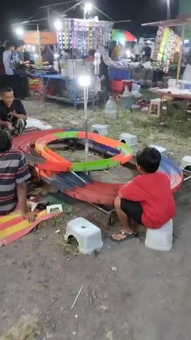 The video captures an outdoor scene at what appears to be a market or fair. In the foreground, a colorful miniature race track is set up. Several people are gathered around it, seemingly engaged in a game or activity involving the track. A young boy in a red shirt is seated on a white stool, looking intently at the track, with a remote control in his hands. To his left, another person, wearing a striped shirt, is also seated on the ground, facing the track. In the background, other individuals and stalls suggest a lively, bustling environment. The lighting indicates it might be evening or under artificial illumination.