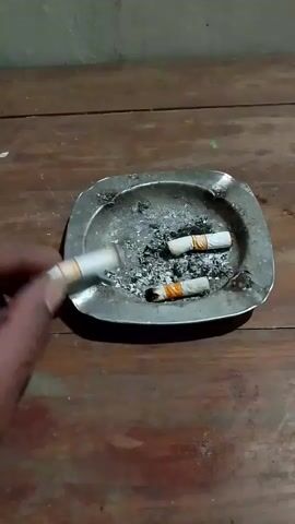 A person's hand holds a lit cigarette over an ashtray on a wooden surface. The ashtray contains cigarette ash and two partially smoked cigarettes with orange bands around their filters. The scene is lit from above, casting shadows.