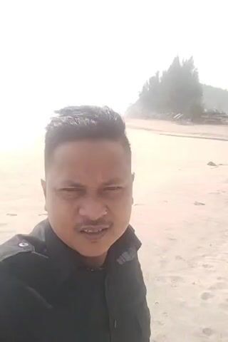 A close-up selfie video shows a man with dark hair and a mustache smiling at the camera. He is wearing a black collared shirt. The background is a beach with sand and a body of water. The sun is bright, creating an overexposed effect. There are some trees and debris visible in the distance.
