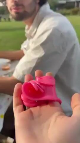 A person holds a pink plastic toy in their hand. The background is blurry, but a man with curly hair and a beard is sitting at a table with a polka dot tablecloth. There are small cups on the table.  The scene appears to be outdoors, with green grass, trees, and a building visible in the distance. The man looks at the camera.