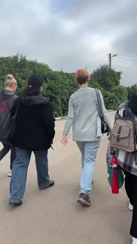 Four individuals are walking away from the camera on a paved road. The person closest to the camera on the right is wearing a plaid backpack. The person next to them is wearing a light blue sweater and light blue pants, carrying a white bag and a gift bag. To their left, a person in a black jacket and blue jeans is walking. The person furthest to the left is wearing a dark grey hooded sweatshirt and black leggings. The road is lined with green bushes and trees. The sky is overcast.