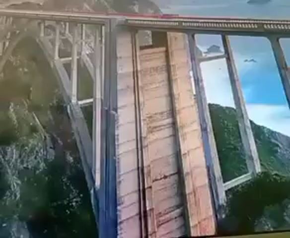 The video shows a close-up shot of a bridge with large arched supports. The bridge is made of concrete and wood. In the background, there are mountains and the ocean. The sky is blue with some clouds. A train is visible on top of the bridge. The camera is panning from left to right, revealing more of the bridge and the surrounding landscape. The overall impression is one of grandeur and a sense of adventure.