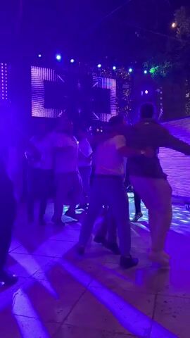 A group of men are dancing in a courtyard at night. They are illuminated by purple and pink lights from a large screen in the background that displays geometric patterns. The men are dressed in casual clothing. Some are wearing white shirts and khaki pants, while others are in darker shirts and pants. They appear to be enjoying themselves, with smiles and energetic movements. The environment suggests an outdoor event, possibly a party or celebration, with trees and string lights visible in the background.