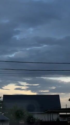 The video shows a landscape shot of a neighborhood in what appears to be the evening. Above the rooftops, power lines stretch horizontally across the frame. A layer of dark, heavy clouds fills the upper portion of the sky, suggesting an overcast or stormy weather. Below these clouds, lighter, scattered clouds are illuminated by a faint, golden light, possibly from the setting sun just beyond the horizon. In the foreground, the silhouette of a house with a dark roof is visible, partially obscured by greenery. The vegetation appears to be a mix of trees and shrubs. Overall, the video presents a scene of a residential area under a dramatic, darkening sky.
