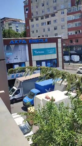 The video opens with a close-up view of a building facade, featuring a prominent sign that reads "AKFA ENERJİ" and "Turkuaz CeraStyle." Below the signs, there are images of water heaters. In front of the building, several vehicles are parked, including a white delivery van, a blue van, and a white truck. Further down the street, more vehicles are visible, including cars and white vans. The scene appears to be an urban environment on a sunny day, with trees and other buildings in the background. The camera then pans to the right, revealing more of the street and a large pine tree in the foreground, obscuring some of the view. The video ends with a wider shot of the street and buildings.