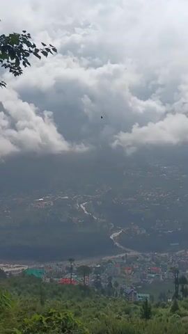 A bird flies through the sky above a valley. The valley contains a town with buildings and a river. The sky is filled with clouds.