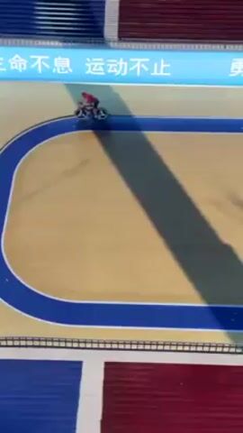 The video shows a cyclist riding on an indoor velodrome track. The track is made of wood and is surrounded by blue and red sections. There is Chinese writing along the top of the track. The cyclist is wearing red and riding a black and blue bike. The cyclist is the only person visible in the video. The cyclist is riding on the blue section of the track on one of the turns.
