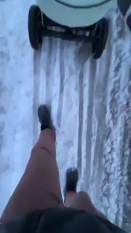 A first-person-view shot shows a person walking in the snow while pushing a baby stroller. The person is wearing shorts and black boots. The ground is covered in snow with tire tracks and footprints visible. The stroller is light blue and gray. The person is walking forward on the snow-covered path.