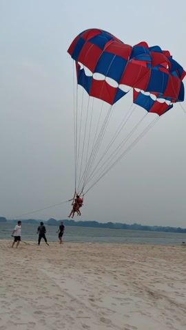 On a beach, under an overcast sky, four people are standing by a parasailer. The parasailer has a large red and blue parachute attached to it. Three people are holding on to the parasailer, helping the two people in the parasailer dismount. The fourth person is standing further away with what appears to be a wooden stick in their right hand. In the background is the ocean, with multiple small land masses. A boat is visible further out. The sand is light colored.