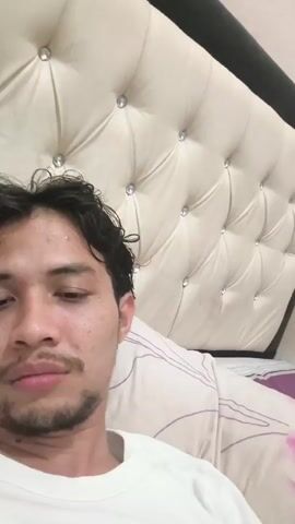 A close-up, low-angle shot shows a young man lying down in bed. He has dark, curly hair and a light beard and mustache. He is wearing a white t-shirt. The background is a large, tufted headboard with diamond-like embellishments. The lighting is dim.
