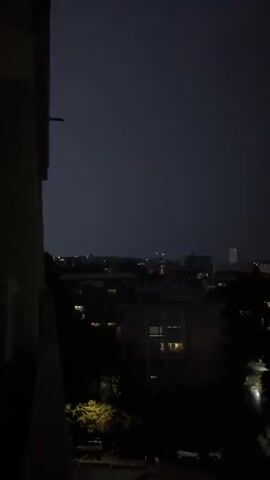 A nighttime view of a cityscape from a balcony. The sky is dark with faint lights from buildings and streetlights scattered across the horizon. In the foreground, a dark silhouette of a building is visible on the left side, and trees with some illuminated leaves are on the right.
