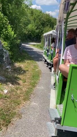 The video begins with a side view of a green and white open-air tram moving along a gravel path. The tram is carrying several passengers, mostly obscured from view. The camera is positioned next to the tram, showing the rear portion of one of the carriages. A young woman in a pink t-shirt with the words "New York" is visible on the right side of the frame. To her left, a young person wearing sunglasses and a dark shirt is seated, followed by other passengers further down the tram, including a person wearing a light-colored hat. The environment appears to be a natural setting with trees and vegetation on the left side of the path, and a stone wall bordering the grass. The sky is bright blue with scattered clouds. The tram continues to move forward, revealing more of the passengers and the path ahead, which curves to the left and disappears into the trees. The lighting suggests it is daytime, likely late morning or afternoon.