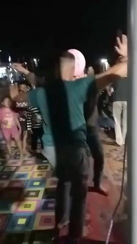 The video shows a nighttime outdoor gathering with several people. In the foreground, two individuals are dancing, with one person in a teal t-shirt and gray pants and another in a dark shirt. A pink balloon is visible between them. To the left, a child in a pink dress is also dancing, and other children are present, some wearing striped shirts. The ground is covered with a colorful, patterned mat. In the background, there are lights and structures, possibly tents or buildings, suggesting a festive event. The overall atmosphere appears to be lively and celebratory.