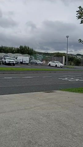 The video shows a parking lot with several cars and vans parked. The sky is cloudy and the weather appears to be overcast. There is a road in front of the parking lot with a pedestrian crossing. There is also a wall with graffiti in the background. The video pans from left to right, showing different parts of the parking lot.