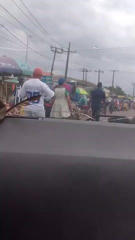 The video shows a street scene with several people visible. In the foreground, part of a car's interior is visible, suggesting the footage is taken from inside a vehicle. The background depicts a busy street with various individuals, some standing, others possibly walking or engaged in activities. There are stalls or shops lining the street, indicated by umbrellas and structures. Power lines and poles are prominent in the background, typical of an outdoor urban or semi-urban environment. The lighting suggests it is daytime, possibly overcast. A few motorcycles are also visible, implying vehicular traffic. The people are dressed in casual clothing, and the overall impression is of a lively, public gathering or marketplace.