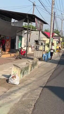 The video captures a street scene in what appears to be a warm, sunny climate. The foreground shows a concrete drainage ditch running alongside a paved road. On the left side of the road, there are several small shops or stalls with awnings providing shade. One stall has a red scooter parked in front of it. A man is standing on the right side of the road, near a cart. He is wearing a white t-shirt and dark shorts, and appears to be looking at something in his hand. Further down the street, other people and vehicles are visible, including a yellow cart being pushed by someone. Utility poles with numerous wires stretch across the sky, indicating an urban or semi-urban environment. The overall impression is of a typical day in a busy street, likely in Southeast Asia, given the architectural style and the presence of scooters.