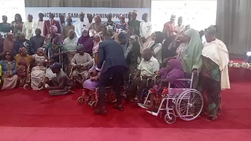 A group of approximately 50 people, mostly seated or in wheelchairs, pose for a photo on a red carpet.  Behind them, a banner reads "Inclusive Family Planning Project".  A man in a suit walks forward to take the picture with his phone, and another person bends down to get out of the shot.  The group is diverse in age and attire, with some wearing traditional clothing and head coverings.  The setting appears to be a large hall with white walls and a high ceiling with some window fixtures.