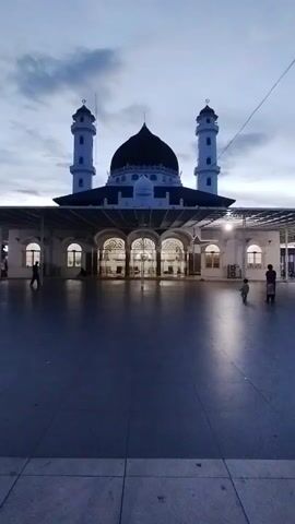 The video shows a mosque at dusk. The sky is cloudy and the light is dim. The mosque has a large dome and two tall minarets. The courtyard in front of the mosque is paved with stone tiles and is mostly empty, except for a few people walking around. The entrance to the mosque is visible, with arched doorways and windows. The overall impression is one of peace and tranquility.