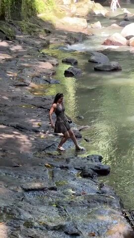 The video captures a daytime scene of a natural river environment in Gianyar, Indonesia. In the foreground, a woman with long dark hair, wearing a dark dress and light-colored sandals, is seen walking cautiously along the rocky bank of a river. She navigates the wet, dark rocks that form the river's edge.

The river itself flows with greenish-brown water, interspersed with numerous large rocks, some partially submerged, creating small ripples or gentle rapids. Lush green foliage lines the banks, indicating a tropical or forested setting. In the background, further upstream, several other people are visible; one appears to be in the water, possibly wading or swimming, while another is on the far bank. Sunlight filters through the overhead canopy, casting dappled light and shadows across the rocky terrain. A small, dark wooden platform or structure is partially visible on the lower right side of the frame. No text is visible in the video.