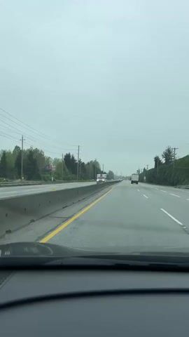 The video provides a first-person view from inside a car driving on a wet, multi-lane highway in Pitt Meadows, Canada, on an overcast day. A concrete barrier separates the opposing directions of traffic. An ambulance with its lights flashing and siren sounding is seen driving past in the opposite direction on the other side of the barrier. Other vehicles, including a white car and a distant truck, are also visible on the highway. Trees, utility poles, and power lines line the sides of the road under a grey sky. No readable text is visible.