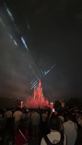 The video shows a nighttime scene of a castle with fireworks and light projections. A crowd of people is gathered in front of the castle, facing away from the camera. The castle is illuminated with red and blue lights, and fireworks are exploding in the sky above it. The people in the crowd are holding up their phones to record the event. The sky is dark and cloudy.