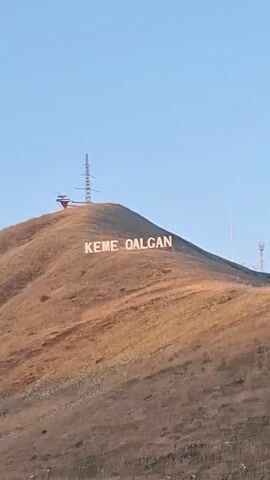 The video shows a sign on a hill that says KEME QALQAN. There are also telecommunication towers on the hill. The hill is covered in dry grass and some small white flowers. The sky is clear and blue. There is a road in the foreground with a guardrail. The video is taken from a moving vehicle.