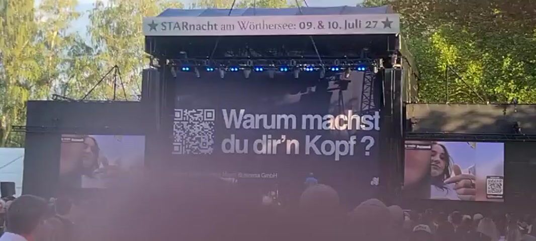 A large banner is displayed on a stage with a QR code and text that reads "Warum machst du dir'n Kopf?". There are screens on either side of the banner, showing images of people. A band is performing on stage. The audience is visible in the foreground. The event is called "STARnacht am Wörthersee".