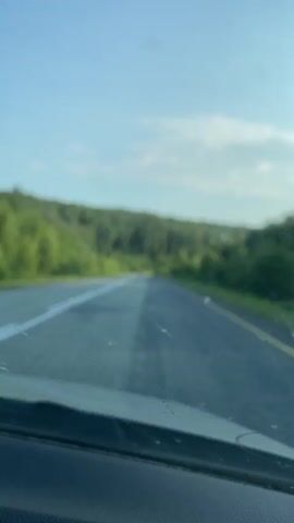 The video shows a view from the front of a vehicle driving on a paved road through a wooded area. The sky is blue with a few scattered clouds. The road has two lanes in the direction of travel, marked with white lines. On the right side of the road, there is a yellow line. The area on either side of the road is covered with green trees and some grassy slopes. In the distance, on the road, there appears to be a dark-colored vehicle. The dashboard of the vehicle is visible in the foreground. The overall impression is that of a scenic drive on a clear day.