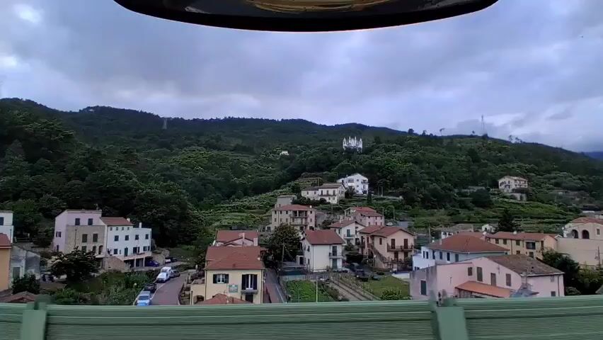 The video offers a scenic view from a moving vehicle, likely a train, looking out over a hillside village in Orco Feglino, Italy. The camera pans slightly and then zooms in on a prominent building.

In the immediate foreground, the top edge of the frame shows a dark, curved part of the vehicle's interior, while a green barrier or railing is visible at the bottom. The midground features a cluster of houses typical of an Italian village, with two to three-story buildings painted in pastel shades like pale yellow, light pink, and beige, all topped with distinct red-tiled roofs. A winding road is visible among the houses, with a few parked cars, and hints of terraced gardens or plots.

The background is dominated by steep, densely forested hills covered in lush green trees. High up on one of these hills, a striking white building with multiple spires or towers stands out against the dark foliage. The camera zooms in on this structure, revealing its ornate, possibly Gothic or neo-Gothic architectural style, resembling a church or sanctuary.

The sky is uniformly overcast with grey clouds, indicating a cloudy day. No people are visible in the scene, and there is no discernible text. The ambient sound of a vehicle's movement, including a mechanical rumble, accompanies the visual. The short clip captures a tranquil landscape observed from a journey.