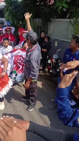 The video shows a group of people participating in a parade, likely a celebration. The participants are predominantly dressed in red and white, common colors associated with national flags. Many are wearing hats, some are holding a large banner with red and white designs. A woman in a red and white outfit, including a festive skirt, is dancing in the foreground. A man in a grey shirt and dark pants with a cap is seen with his arm raised, possibly waving or directing. Several children are visible in the background, also dressed in red and white. A woman is holding a baby. The environment appears to be a street lined with houses and trees, with some motorcycles parked along the side. The overall atmosphere seems festive and energetic.