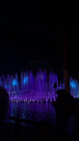 At night, a crowd watches a fountain show of water shooting into the air lit with purple light. The Pixar Pal-A-Round Ferris wheel is illuminated in the distance. The show takes place on a lake surrounded by decorative railings. A loudspeaker and pole are visible in the foreground.