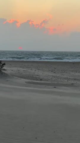 The video shows a beach scene at sunset. The sky is orange and pink, with a few clouds. The sun is a red disc on the horizon, partially obscured by the clouds. The ocean is rough, with waves crashing on the shore. The beach is sandy, with some windblown sand creating ripples on the surface. A person is walking away from the camera towards the ocean. They are wearing a pink shirt and black shorts. They have a bag slung over their shoulder. The person's hair is short and blonde.