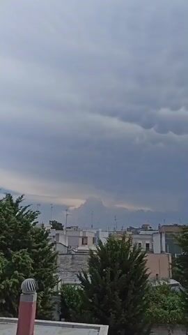 The video shows a view of the sky above a town. The sky is filled with dark, ominous clouds, some of which have a distinctive mammatus appearance, resembling a cluster of bubbles or pouches hanging from the underside of a cloud. In the distance, a larger cloud formation is visible, possibly a cumulonimbus cloud, with a lighter area near its base, suggesting a potential storm. Below the clouds, the rooftops of buildings are visible, with antennae protruding from some of them. Trees with dense green foliage frame the foreground and sides of the shot, obscuring parts of the town. The overall atmosphere is moody and suggests impending bad weather.