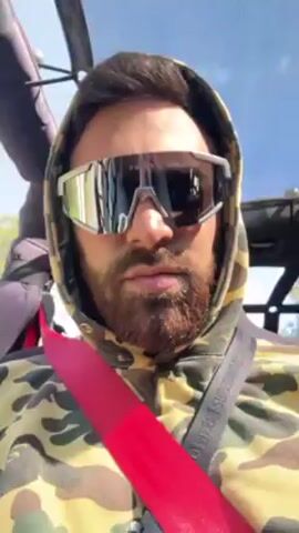 A man with a beard and wearing sunglasses and a camouflage hoodie is sitting in a vehicle, possibly an off-road vehicle, with a red seatbelt fastened. The camera is positioned close to the man, capturing a close-up view of his face. The man is looking directly at the camera with a neutral expression. The background is blurry but appears to show trees and sky, suggesting the vehicle is moving outdoors.
