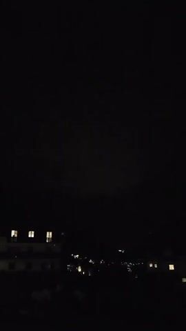 The video shows a dark sky over a dimly lit town at night. A few windows in buildings are illuminated. A faint, diffused light source is visible in the sky, possibly lightning or a distant light.